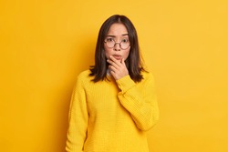 Portrait of worried surprised Asian woman holds chin looks concerned at camera wears transparent glasses and sweater isolated over yellow background. People face expressions and reaction concept