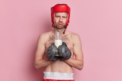 Shirtless male boxer has displeased face expression holds bottle of milk wears sport gloves feels tired after training isolated over pink background gets good nutrients prepares for important fight