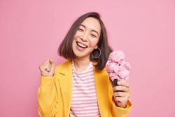 Joyful adorable teenage girl with eastern appearance dark hair tilts head clenches fist eats tasty ice cream smiles broadly has fun enjoys summer dressed in stylish clothes isolated on pink wall