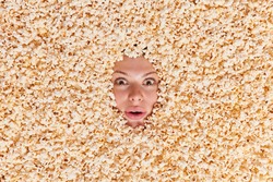 Overhead shot of surprised woman stares bugged eyes feels impressed being fully covered with delicious popcorn. Snack for cinema goers. Female model buried in air popped corn has shocked look