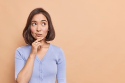 Thoughtful Asian woman keeps hand on chin looks pensively above dressed in casual blue jumper poses against brown background blank copy space for your advertising content thinks about future