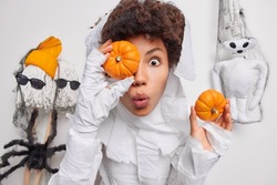 Surprised woman covers eye with orange pumpkin cannot believe her eyes dressed in scarying costume prepares decorations for halloween isolated over white background. Spooky holiday is almost there