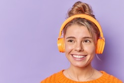 Dreamy lovely young woman smiles broadly enjoys audio track in wireless headphones concentrated away with dreamy expression isolated over purple background blank empty space for your promotion
