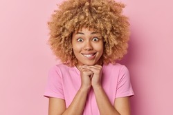 Portrait of surprised young woman with curly bushy hair keeps hands under chin bites lips has wondered expression wears casual t shirt poses against pink background. Human facial expressions