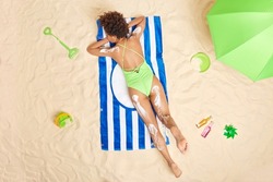 Overhead shot of relaxed slim woman dressed in green swimsuit lies on stomach sunbathes at beach applies skin care cream has lazy day enjoys summer vacation. People leisure relaxation concept