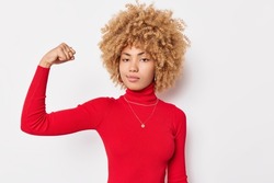 Portrait of self confident curly haired woman flexes biceps shows arm muscles looks seriously at camera wears red turtleneck poses against white background has workout. She can stand up for herself