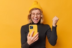 Big luck concept. Overjoyed fair haired young woman clenches fist exclaims from joy exclaims with excitement wears hat and black turtleneck isolated over yellow background won online lottery
