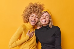 Sincere human emotions concept. Positive overjoyed young women have fun laugh gladfully smile toothily cannot stop laughing stand closely to each other dressed casually isolated over yellow wall