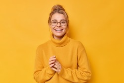 Pretty blonde European woman smiles toothily keeps hands together feels satisfied wears round spectacles and casual loose jumper isolated over vivid yellow background. Happy emotions concept