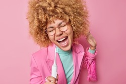 Carefree optimistic woman has fun while brushing teeth holds electric toothbrush near mouth as if microphone sings favorite song carries ladies bag dressed in stylish clothes isolated over pink wall