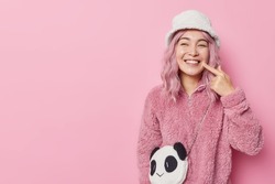 Pretty pink haired Asian woman points at toothy smile points at white teeth wears fashionable outerwear looks gladfully away isolated over pink background blank copy space for your promotion