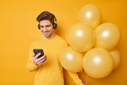 People and holidays concept. Happy adult man checks message via smartphone listens favorite music in headphones wears casual jumper holds bunch of inflated balloons isolated over yellow wall