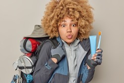 Studio shot of surprised woman with curly hair keeps lips folded holds toothbrush and tube of toothpaste dressed in activewear carries heavy rucksack spends free time on camping isolated on grey wall