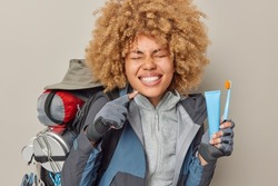 Positive woman points at toothy smile shows white teeth holds toothbrush and toothpaste spends free time actively takes care of oral hygiene dressed in jacket carries rucksack isolated on grey wall