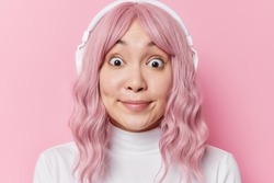 Headshot of young Asian woman with pink hair looks with widely opened eyes feels amazed wears white turtleneck finds out shocking news listens audio track via headphones isolated over rosy wall