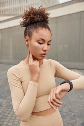 Outdoor shot of serious woman with curly hair gathered in bun checks heart rate focused at smartwatch monitors pulse dressed in activewear leads healthy lifestyle goes in for sport regularly.