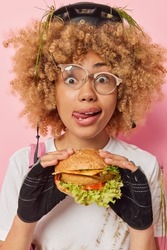 Photo of surprised curly haired woman feels temptation to eat tasty burger after bike travel licks lips with tongue wears protective gear and damages eyeglasses fell off bicycle poses indoors