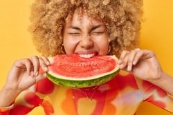 Photo of curly haired young woman eats juicy delicious watermelon bites favorite fruit keeps eyes closed from enjoyment poses against vivid yellow background. Summer time and nutrition concept