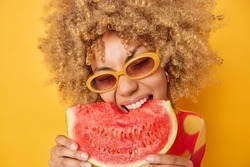 Summer taste. Close up portrait of curly haired young woman bites big red slice of fresh natural watermelon with appetite wears sunglasses likes juicy fresh fruit isolated over yellow background.