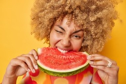 Close up shot of curly haired cheerful woman holds bigs slice of juicy watermelon enjoys eating delicious summer dessert isolated over bright yellow background. Healthy eating lifestyle concept