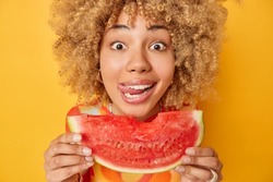 Summer time concept. Close up shot of curly woman holds half of red watermelon licks lips enjoys eating tasty juicy fruit looks surprisingly at camera isolated over bright yellow background.