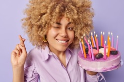 Cheerful birthday girl crosses fingers makes wish before blowing out candles on festive cake winks eye and bites lips hopes dreams come true isolated over purple background. Holiday concept.