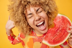 Cheerful beautiful woman with curly bushy hair holds big slice of juicy watermelon keeps mouth opened dressed in casual jumper isolated over vivid yellow background. Summer lifestyle concept