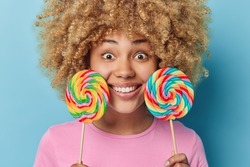 Headshot of positive curly haired woman holds two big multicolored lollipops smiles happily has wondered expression dressed in casual pink t shirt isolated over blue backgroound. Sweet tooth