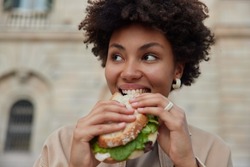 Pretty curly haired woman bites delicious sandwich poses outdoors at street looks away dressed casually has quick snack while walking outside being hungry. People lifestyle and fast food concept