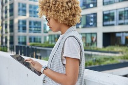 Half length shot of beautiful curly haired woman in elegant formal clothes satisfied with successful career reads text on her digital tablet during recreation time poses against office building