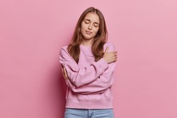 Love yourself concept. Indoor shot of lovely dark haired woman keeps eyes closed embraces herself wears casual jumper and jeans tilts head isolated over pink background. Self acceptance concept