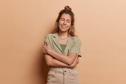 European girl wears shirt trousers and embraces herself with warm smile sstands isolated against beige background emanating sense of inner peace and self acceptance isolated over brown background