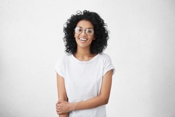 Positive human facial expressions and emotions. Isolated shot of attractive dark-skinned student girl in round eyewear looking and smiling broadly at camera during nice conversation with someone