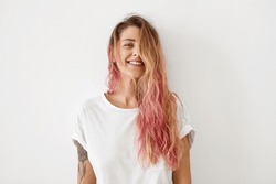 Horizontal portrait of pleasant-looking Caucasian female with long hair, pink on tips, having tattooes on arms, wearing white casual T-shirt, covering her face with hair, looking happily in camera