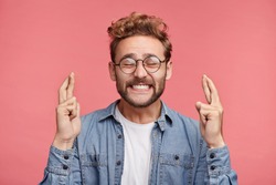 Glad positive bearded man crosses fingers, closes eyes with pleasure, anticipate hearing good news, isolated pink background. Happy male rejoices that his dreams came true. Excitement concept