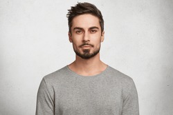 Isolated shot of young handsome male with beard, mustache and trendy hairdo, wears casual grey sweater, has serious expression as listens to interlocutor, poses in studio against white background