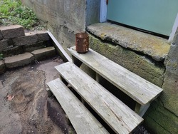 A rusty can sitting on a set of old wooden stairs. The staircase is made of wood and leads up to the cracked foundation of the home that is green with mold and mildew.