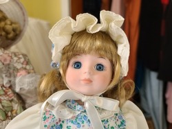 A closeup of the face of a doll. The doll has blue eyes, blond hair, and is wearing a white bonnet with a white ribbon around its face.