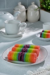 On a white table, there is a large plate of rainbow roll cake filled with chocolate glaze, a white cup, a white teapot and a small plate of rainbow cake ready to eat, there is a shadow on the table 