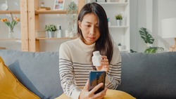 Sick young Asian woman hold medicine sit on couch video call with phone consult with doctor at home. Girl take medicine after doctor order, quarantine at home, Social distancing coronavirus concept.