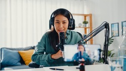 Teenage Asia girl influencer use microphone wear headphone record content with smart phone for online audience listen at house. Student female podcaster make audio podcast from her home studio.