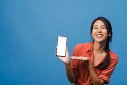 Young Asia lady show empty smartphone screen with positive expression, smiles broadly, dressed in casual clothing feeling happiness on blue background. Mobile phone with white screen in female hand.