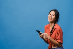 Surprised young Asia lady using mobile phone with positive expression, smile broadly, dressed in casual clothing and looking at camera on blue background. Happy adorable glad woman rejoices success.