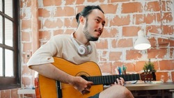 Artists producing music in their home sound studio, Asian man playing guitar and singing in living room at home. Lifestyle man relax in morning at home concept.