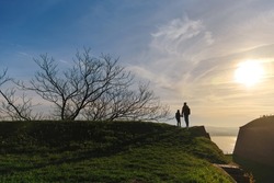 Silhouette of father and son standing on Petrovaradin Fortress fortress hilltop overlooking the Petrovaradin fortress, Danube river and Novi Sad cityscape at sunset. Family travel to Serbia, Balkans.
