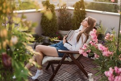 Young romantic woman holding book to her chest on urban rooftop garden at sunset. Dreamy girl reading exciting novel while sitting in chaise longue on cozy terrace with flowers, plants and city view.