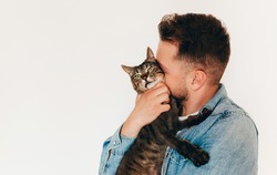 A young man in blue denim shirt is holding and hugging a cute funny brown tabby cat. Home pets. Light background. Pet lover. Copy space for text
