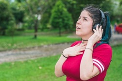 beautiful college woman with blue hair and red dress, listening to music with her wireless headphones in a park, excited and sentimental girl listening to songs with one hand on her chest