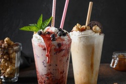 Two delicious berry milkshake garnished with whipped cream, blueberries, dripping jam on dark background. Close up.