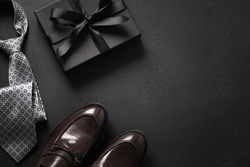 Happy Fathers Day greeting card with gift box. Set of classic mens clothes - brown shoes, tie and gift on black background. Men's accessories set. Top view. Copy space.